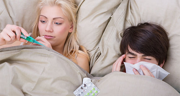 a couple in bed - the woman checking a pregnancy test while the man is blowing his nose with a tissue
