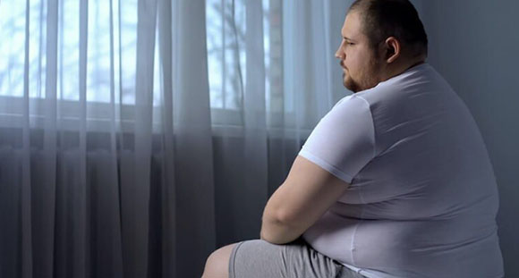 an obese man sitting on the edge of a bed with a sad expression on his brow
