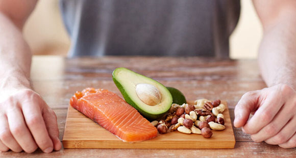 a man sitting in front of a wooden board with a fillet of salmon, half and avocado, and an assortment of nuts