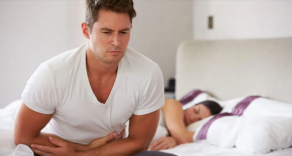a man holding his stomach in pain from Crohn's Disease, with a woman sleeping behind him