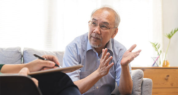 an older Asian man sitting on a sofa speaking with a professional