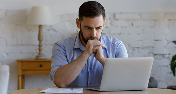 a bearded man sitting at a table looking at a laptop with a pen and paper next to it
