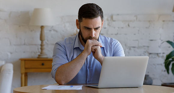 a worried man sitting at a table looking at a laptop