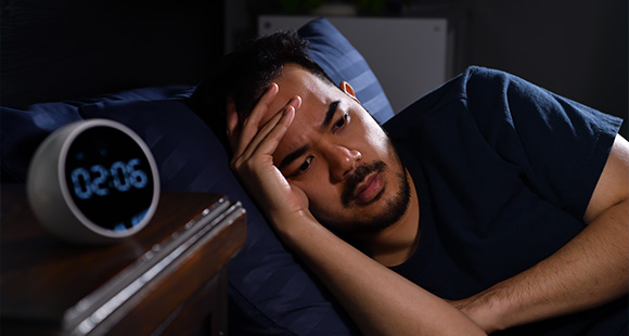 an insomniac man sitting awake in bed next to an alarm clock showing 2AM